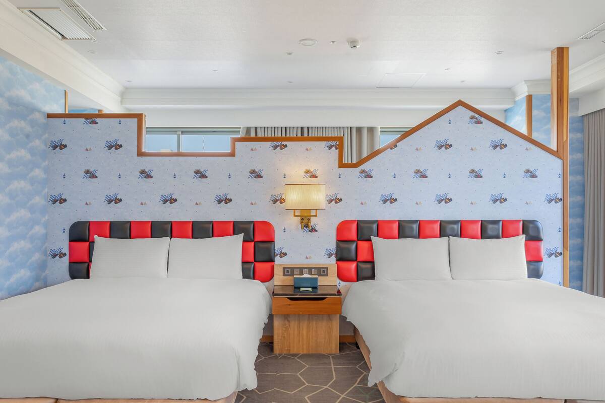 A hotel room with two beds, a wooden nightstand, a wall-mounted lamp, and a window with curtains.