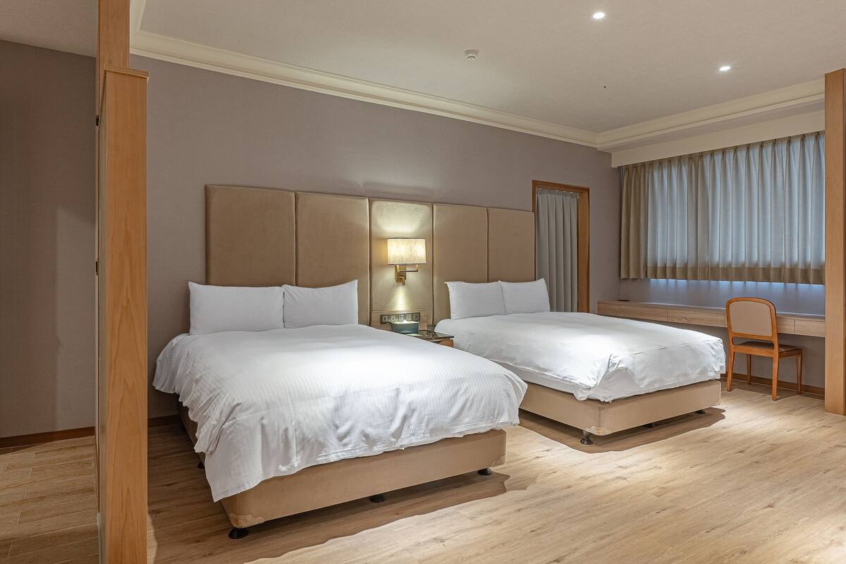 A hotel room with two beds, a desk, a chair, and a window with curtains.