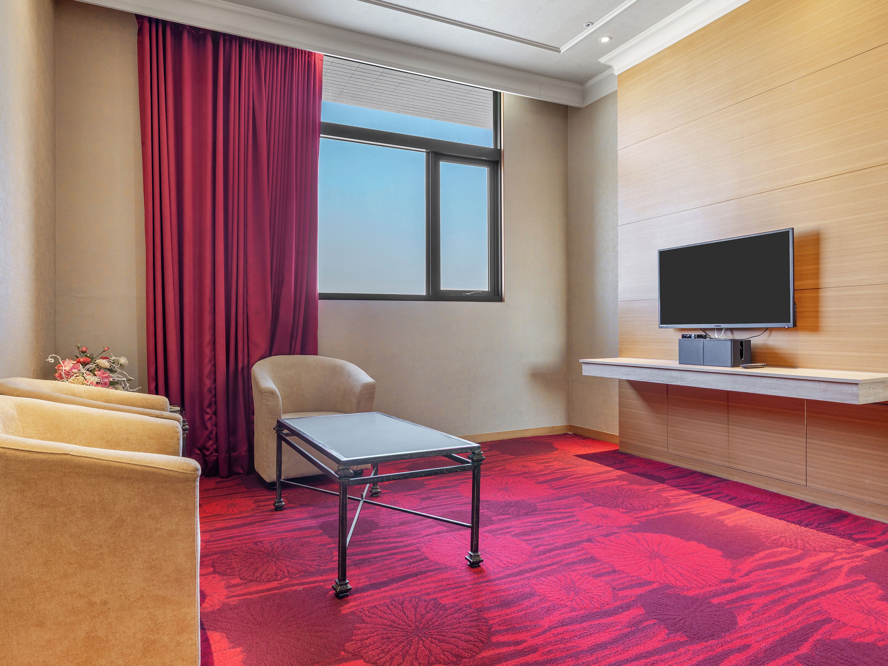 Une chambre d’hôtel avec un tapis rouge, un canapé, une petite table, une chaise, un téléviseur posé sur un meuble et une fenêtre avec des rideaux.
