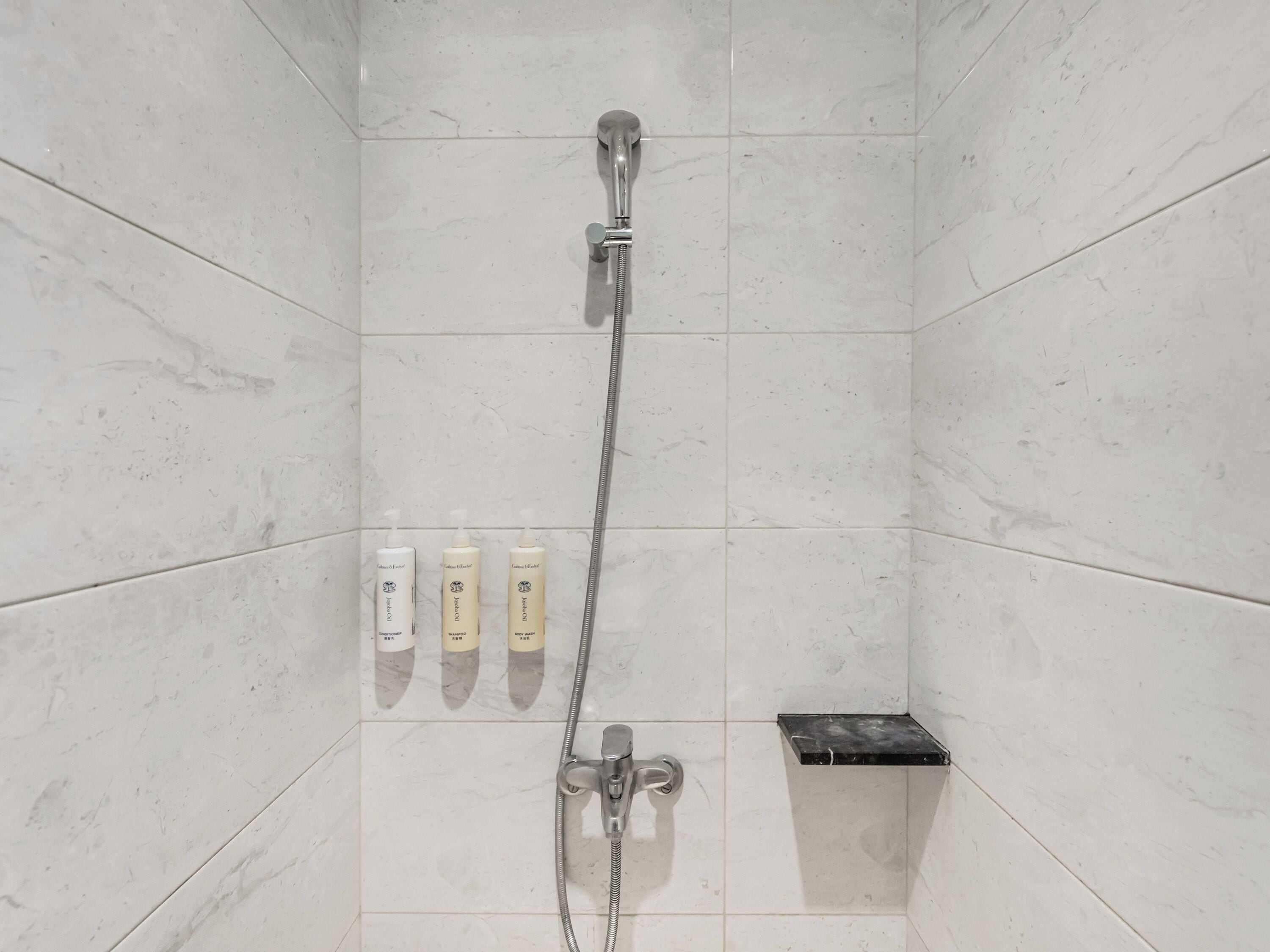 A shower area with a handheld showerhead, a wall-mounted faucet, and three bottles of shampoo on the wall.