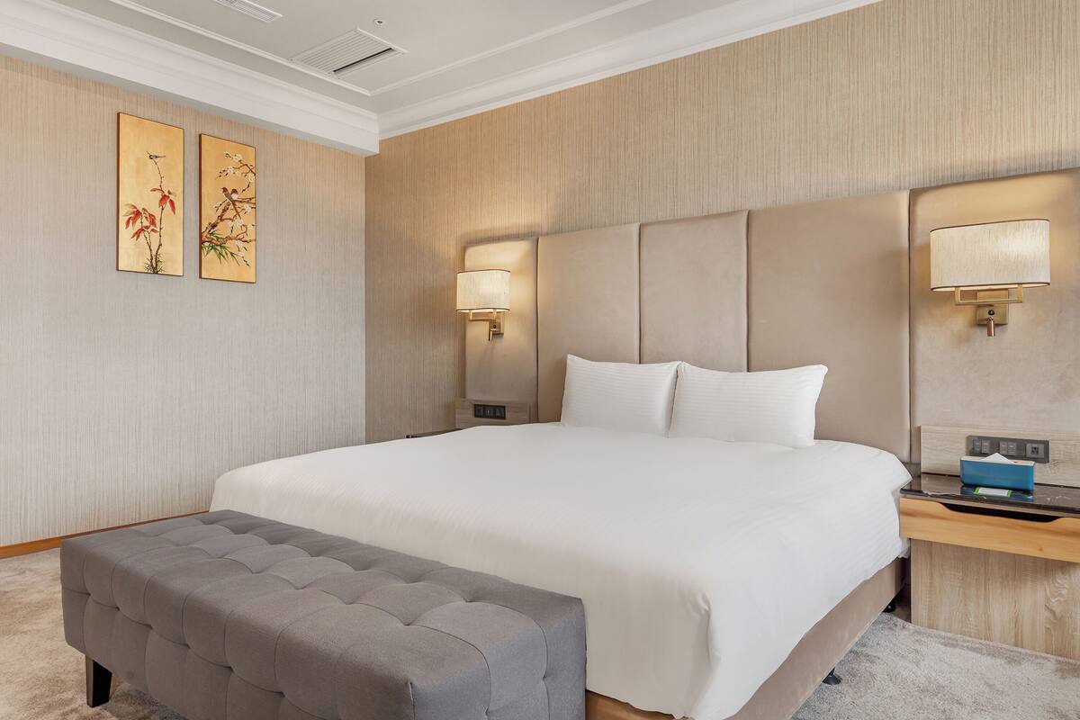 A hotel room with a large bed, two wall-mounted lamps, a bedside table with a lamp, a grey upholstered bench, and two wall art pieces.
