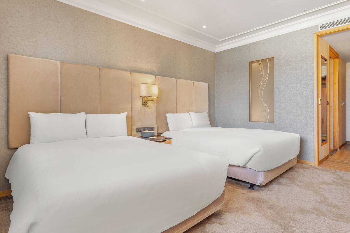 A hotel room with two beds, a nightstand, a wall-mounted lamp, and a framed artwork.