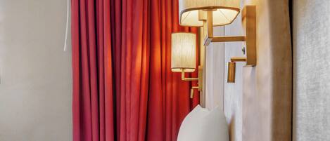 Habitación de hotel con cama, cortinas rojas, lámpara de pared y mesita de noche con caja de pañuelos.