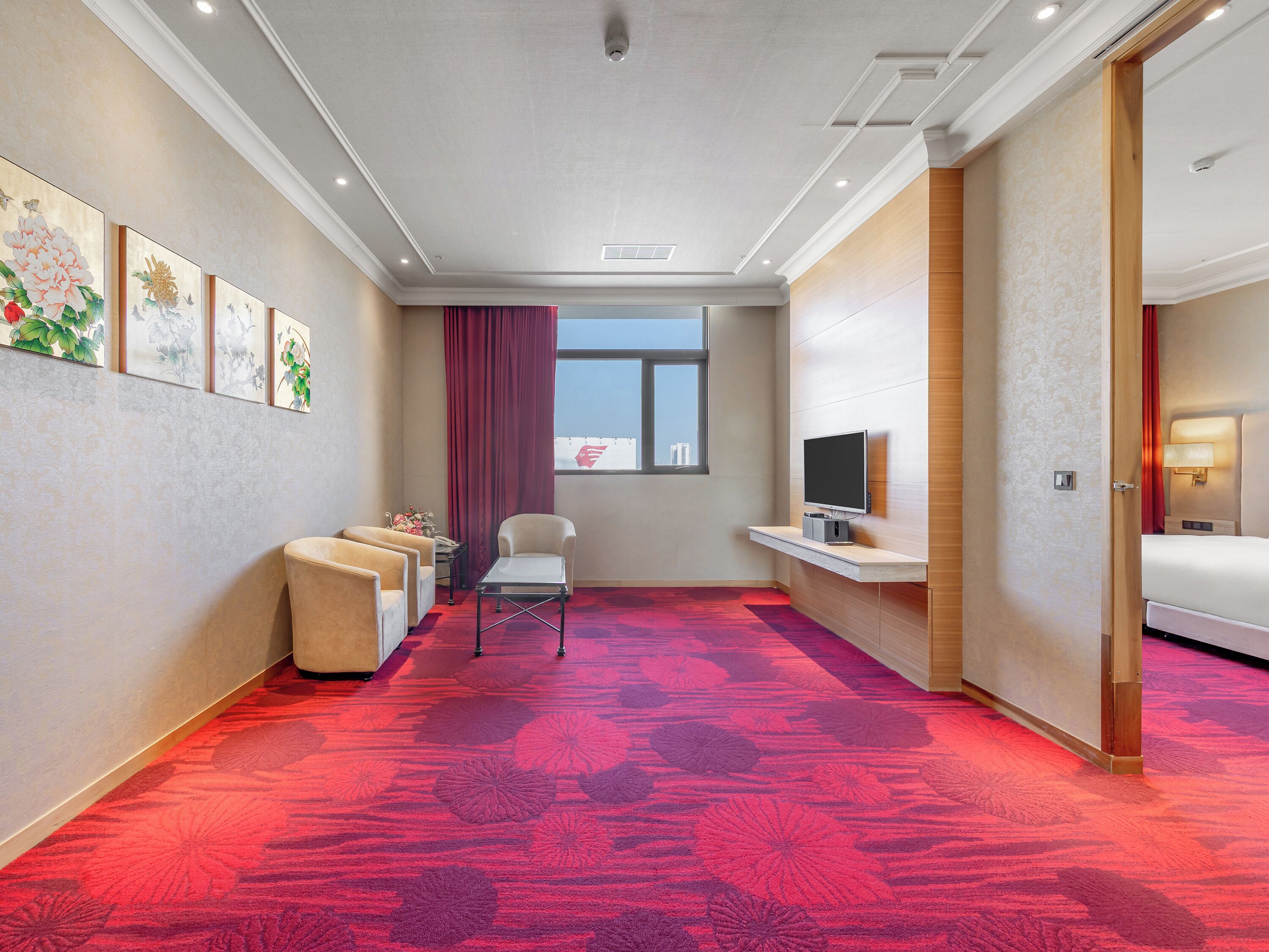 A hotel room with a red carpet, a TV on a stand, a bed, a chair, and artwork on the wall.