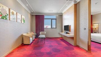 A hotel room with a red carpet, a TV on a stand, a bed, a chair, and artwork on the wall.