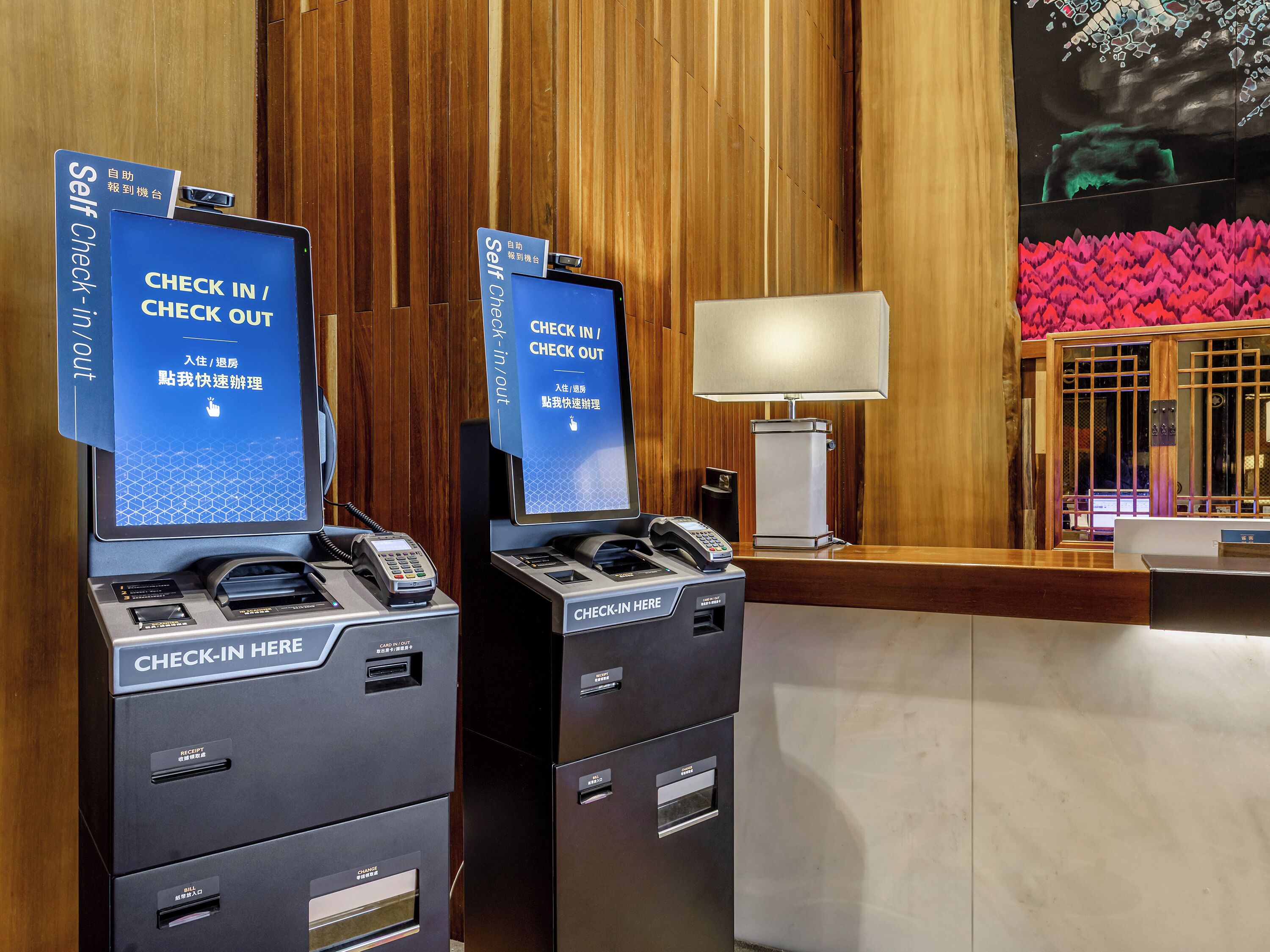 Un espace d’accueil d’hôtel comprenant des bornes d’enregistrement et de départ en libre-service, un comptoir de réception et un mur décoratif.
