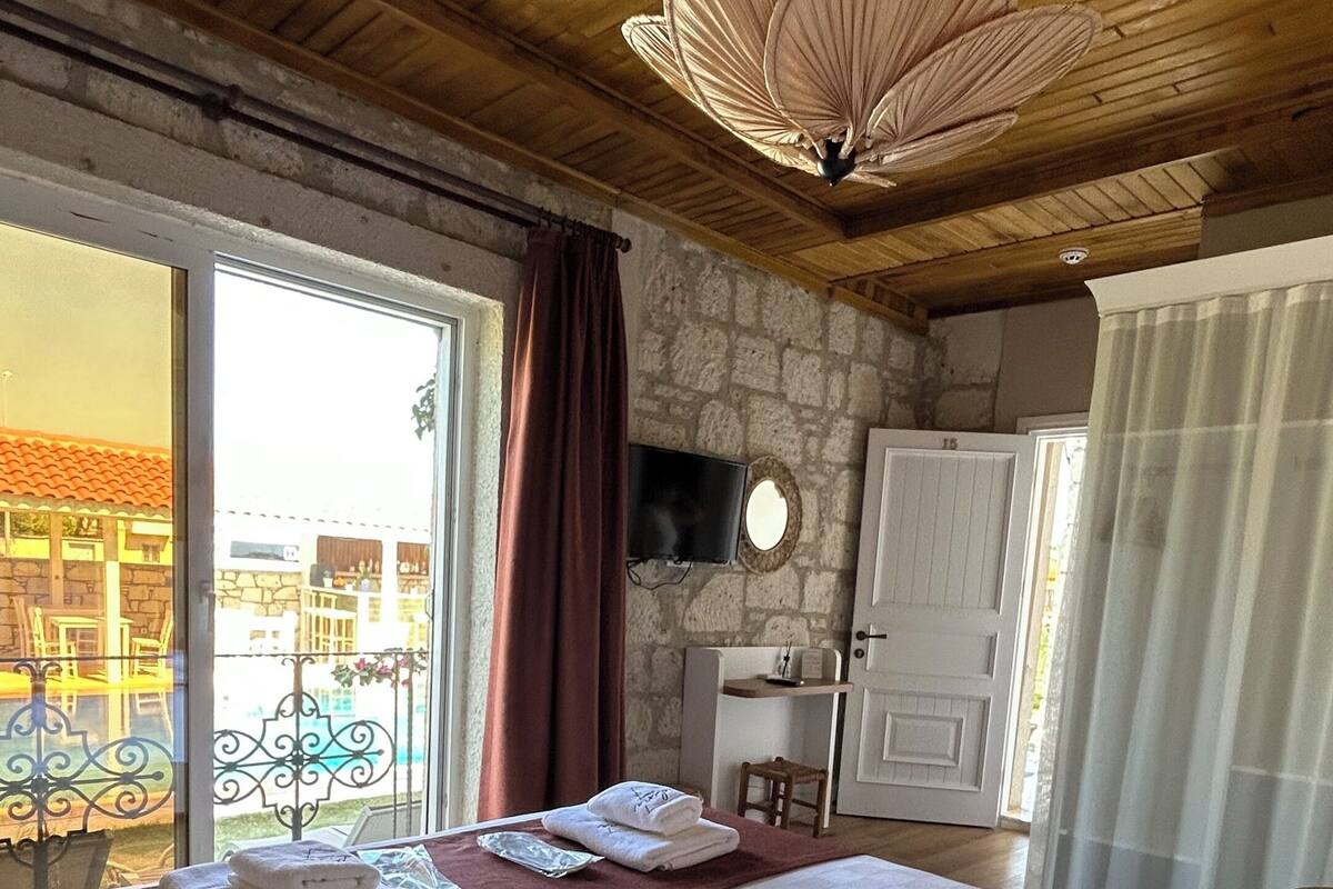 A hotel room with a large bed, a stone wall, a wooden ceiling, a TV, and a view of the outside.