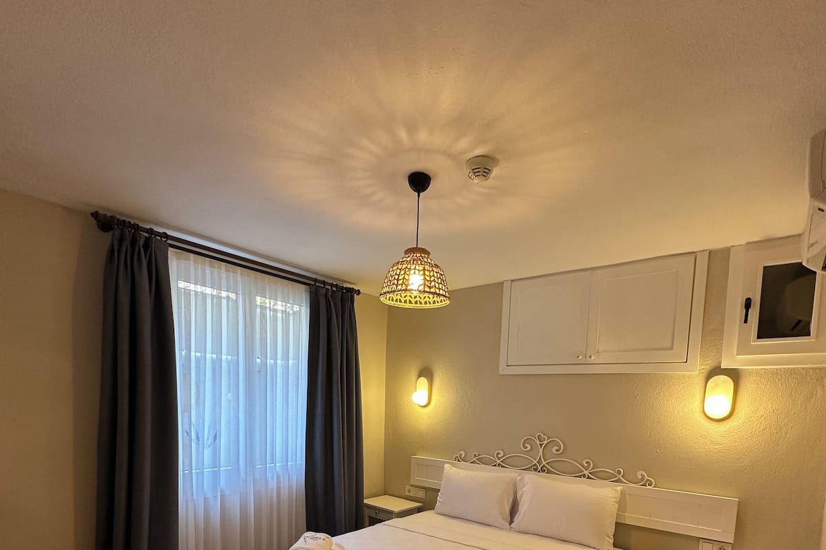 A neatly made bed with towels, a bedside table, a nightstand, and a hanging light fixture in a room with wooden flooring and a window with curtains.