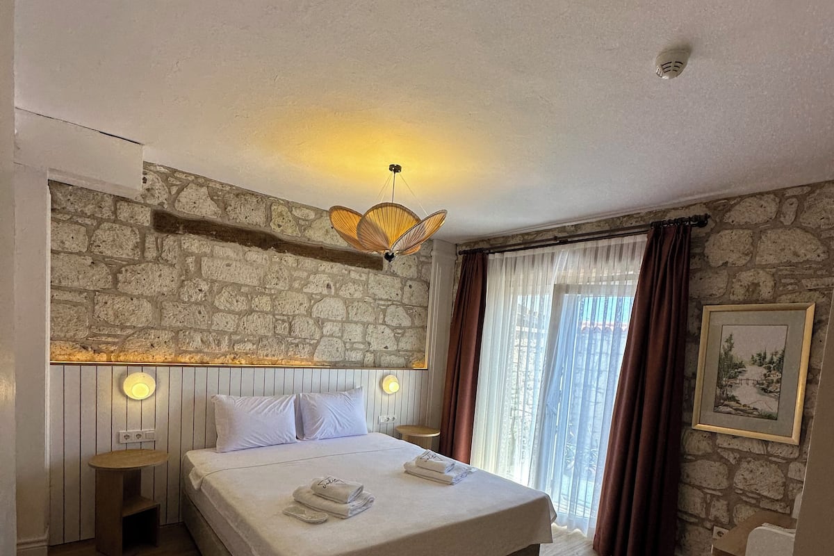 A bedroom with a large bed, a stone wall, a wooden floor, a bedside table, and a hanging light fixture.