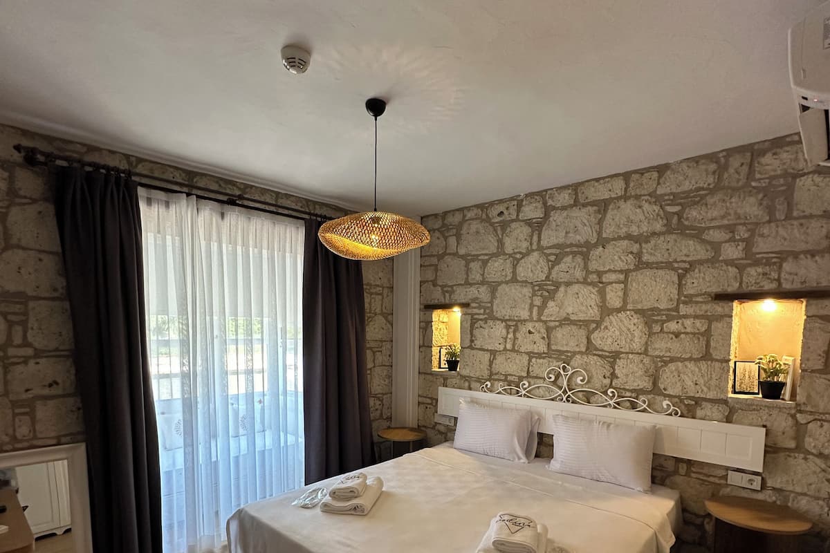 A hotel room with a large bed, stone wall, two bedside lamps, and a view of the outdoors.