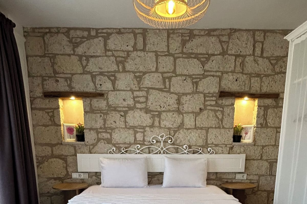A bedroom with a stone wall, a bed with white linens, a hanging light fixture, and two bedside lamps.