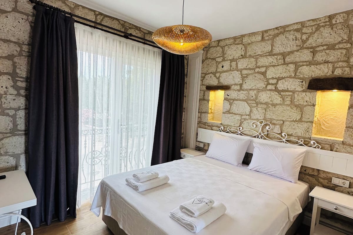 A hotel room with a large bed, a small table, a chair, a stone wall, and a window with curtains.