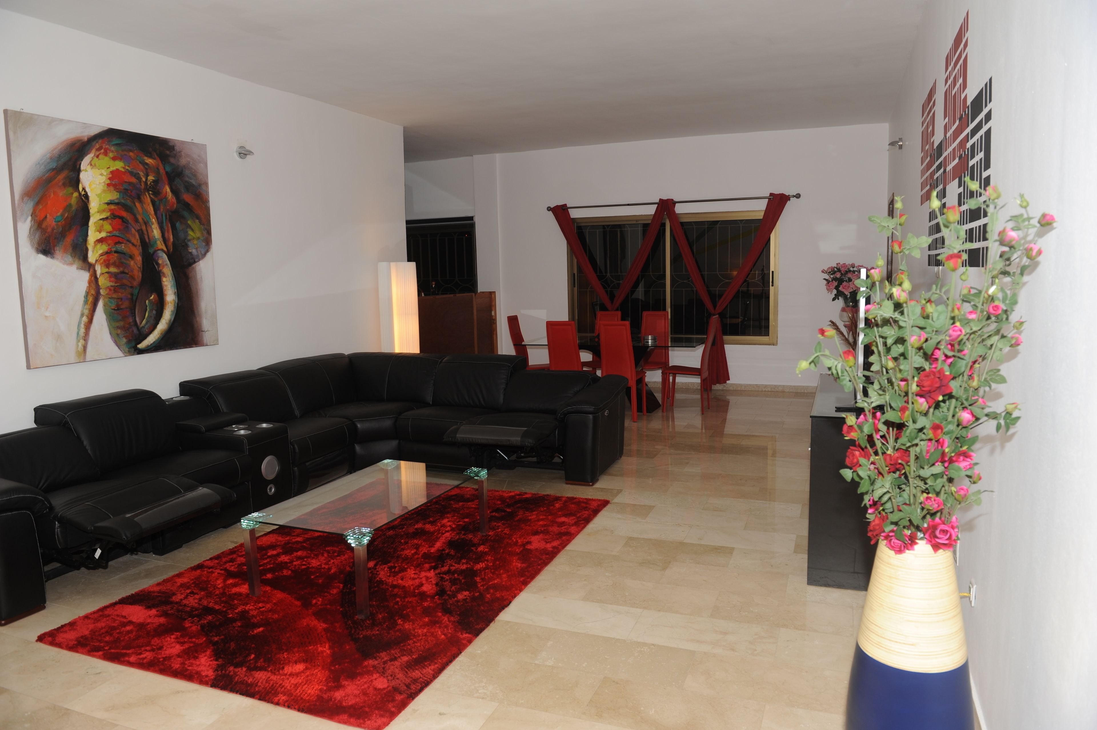 A living room with a black sectional sofa, a red rug, a glass coffee table, and a vase of flowers.
