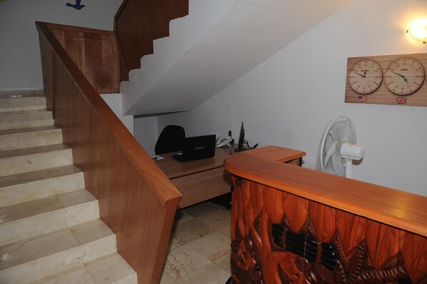 Una escalera con barandillas de madera, un escritorio con una computadora, un ventilador y dos relojes de pared mostrando diferentes husos horarios.