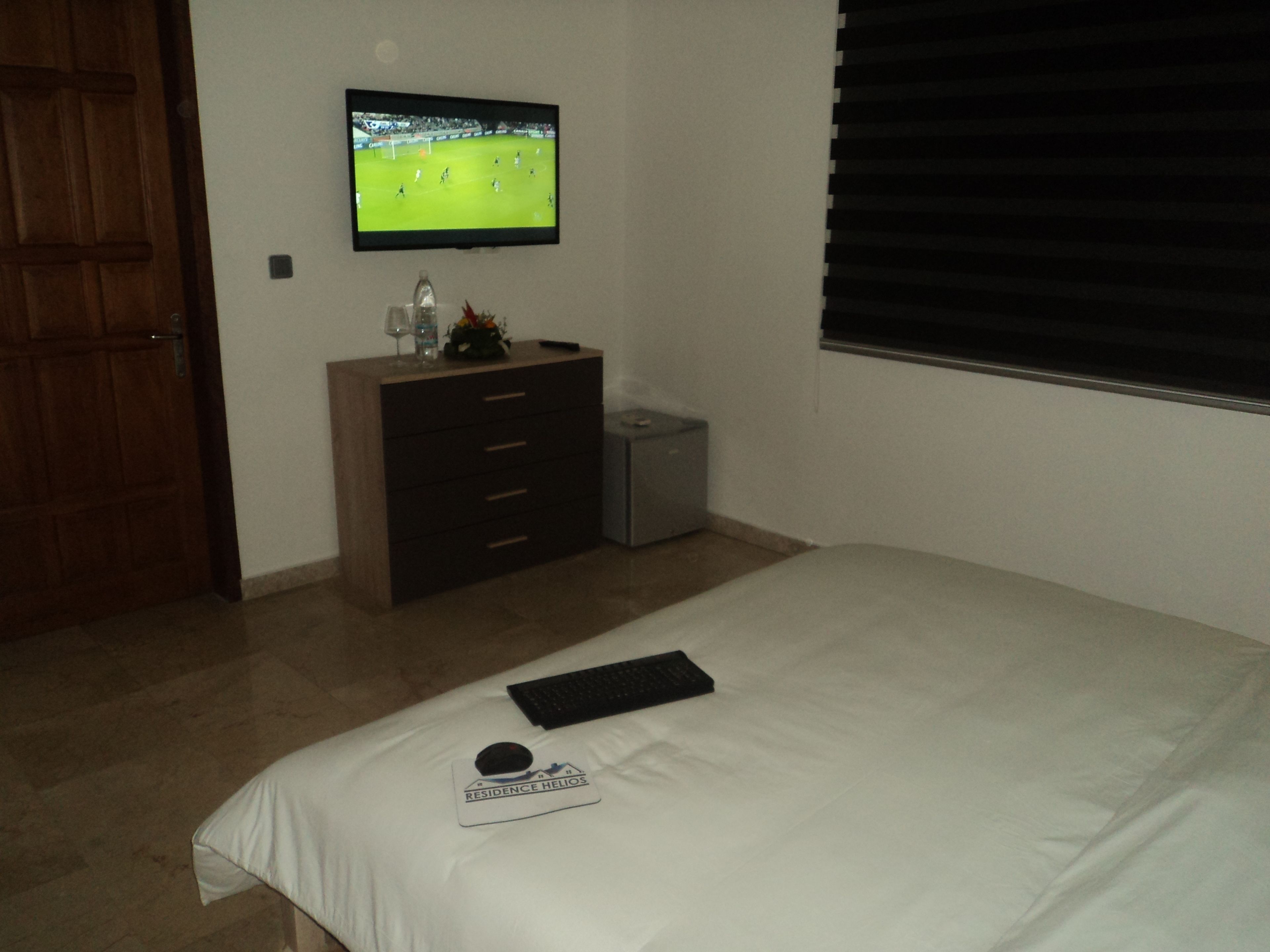 A bedroom with a bed, a TV mounted on the wall, a dresser, and a window with blinds.