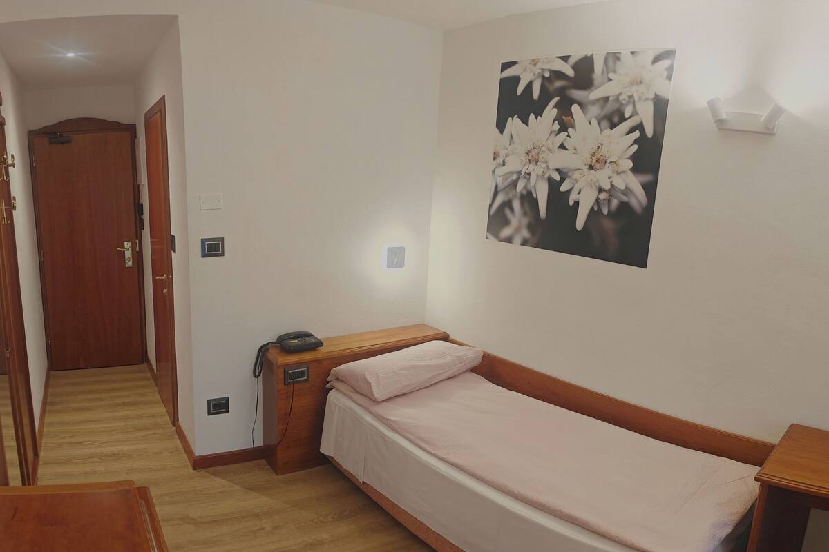 A small, neatly arranged bedroom with a single bed, a bedside table, and a wall-mounted light.
