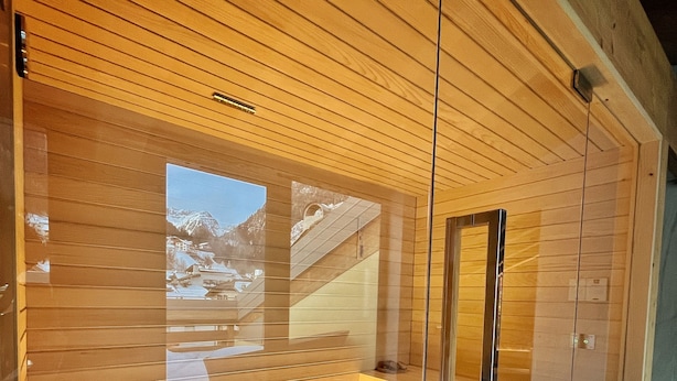 A modern sauna room with wooden slats on the ceiling, a glass door, and a stone bench. - Hotel Moena (Moena)