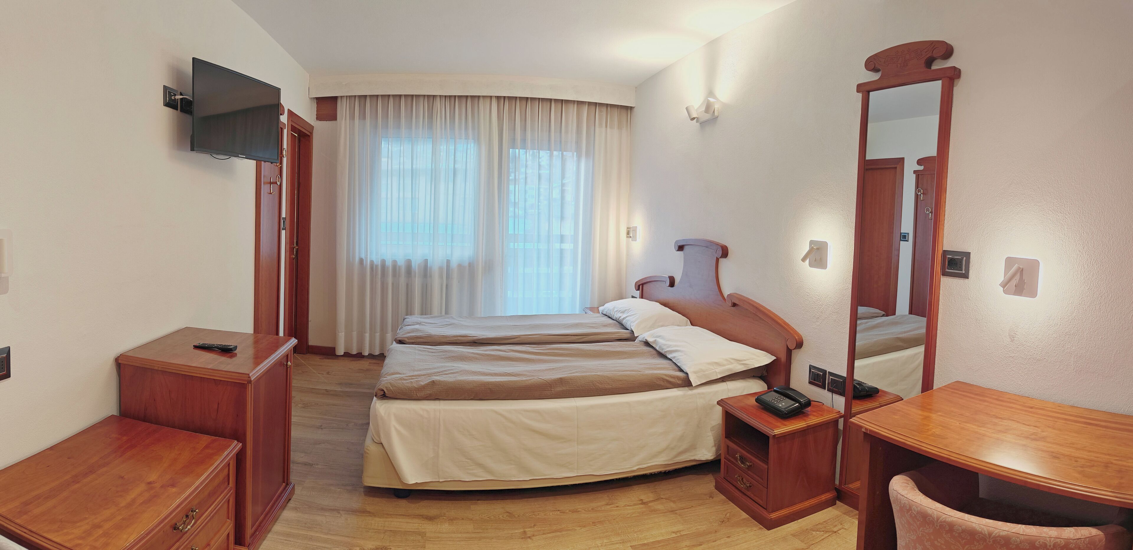 A hotel room with two beds, a TV mounted on the wall, a bedside table with a phone, a mirror, and a wooden nightstand.