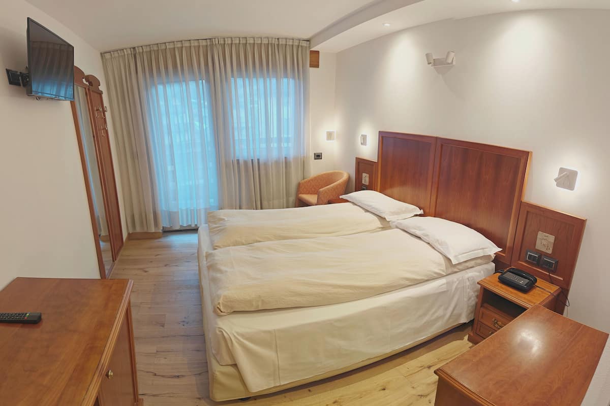 A hotel room with a double bed, a wooden nightstand, a chair, a TV mounted on the wall, and a window with curtains.