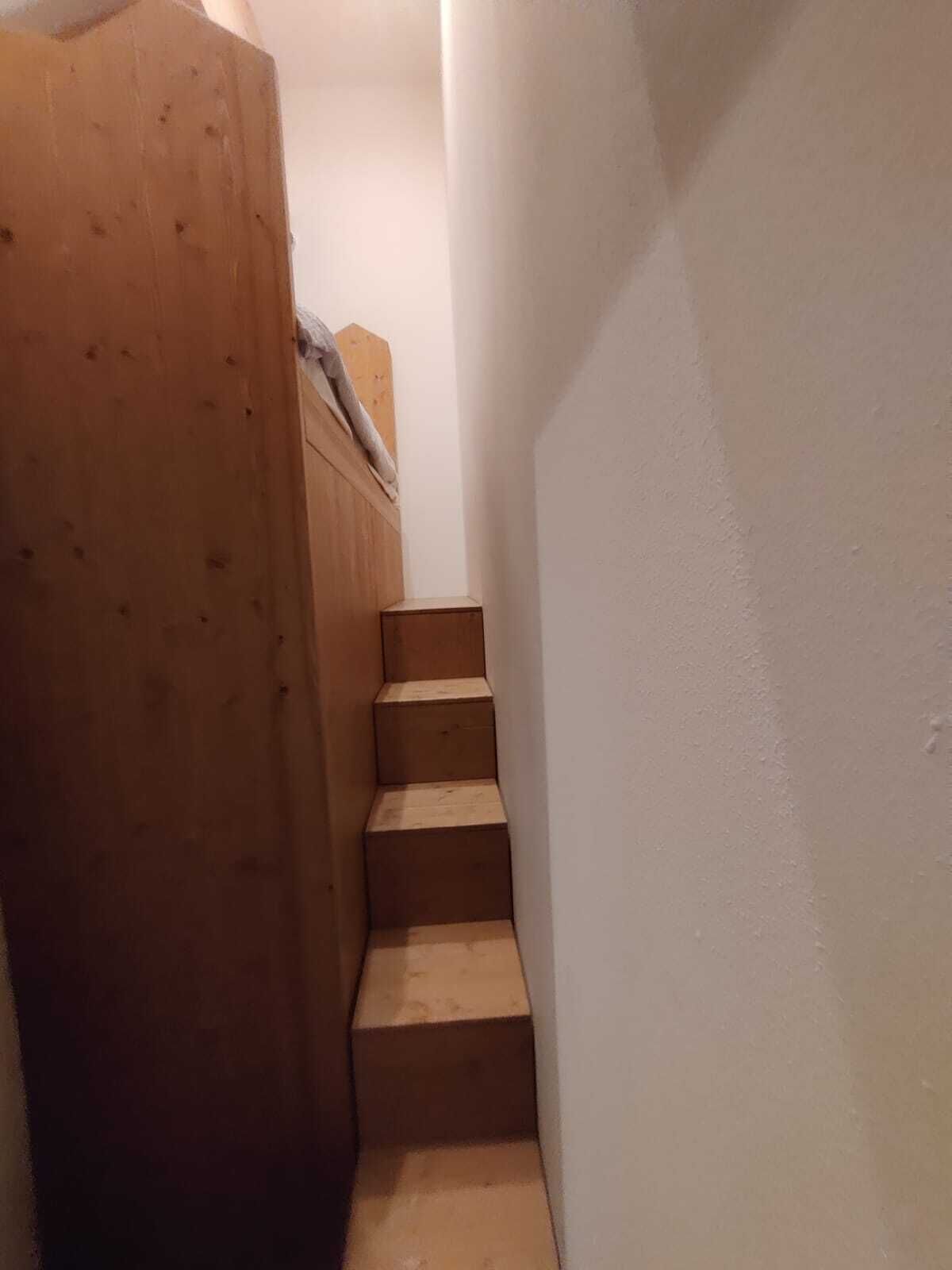Un estrecho pasillo con escalones de madera que suben a una zona con una cama tipo altillo.