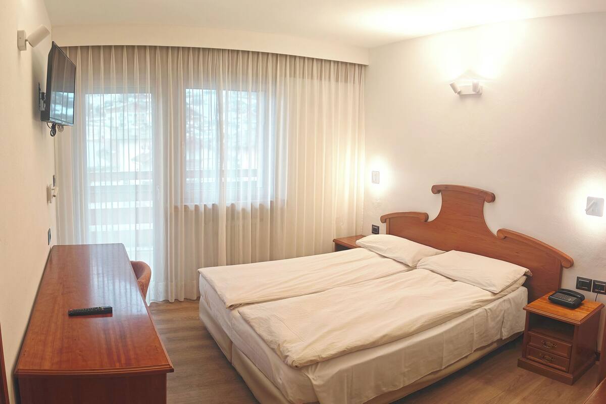 A hotel room with a double bed, a wooden desk, a TV mounted on the wall, and a window with curtains.