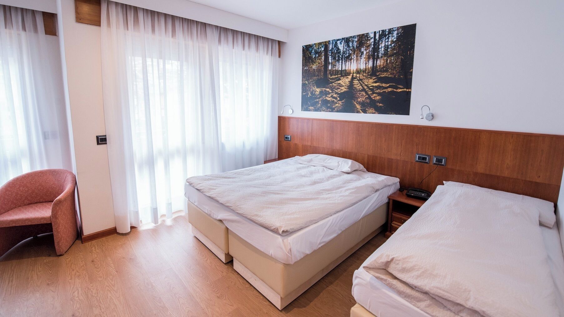A hotel room with a bed, a chair, a bedside table, a painting, and a window with curtains.