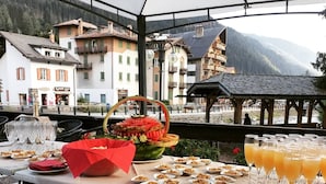 An outdoor buffet setup with various food items and drinks, set against a backdrop of buildings and mountains. - Hotel Moena (Moena)