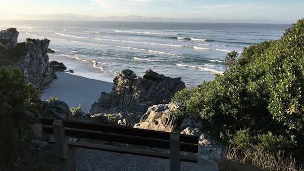 Vista costeira com falésias rochosas, uma praia arenosa e um banco em primeiro plano.