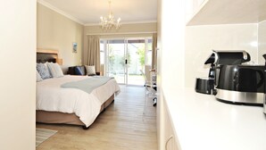 Quarto de hotel com cama, um candelabro, um ventilador de teto, uma cafeteira e vista para o exterior através de uma porta de vidro.