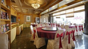 Een balzaal met ronde tafels, witte stoelen met rode linten, een kroonluchter en uitzicht door grote ramen.