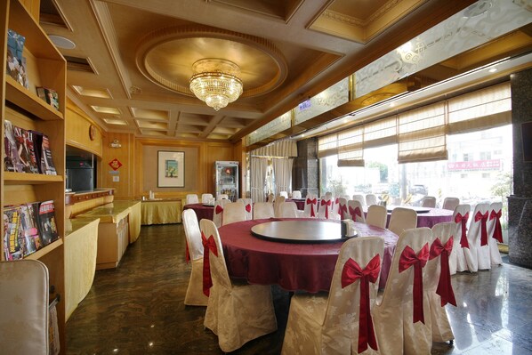 Un salón de banquetes con mesas redondas, sillas blancas con lazos rojos, una araña de cristal y vistas amplias a través de grandes ventanales.