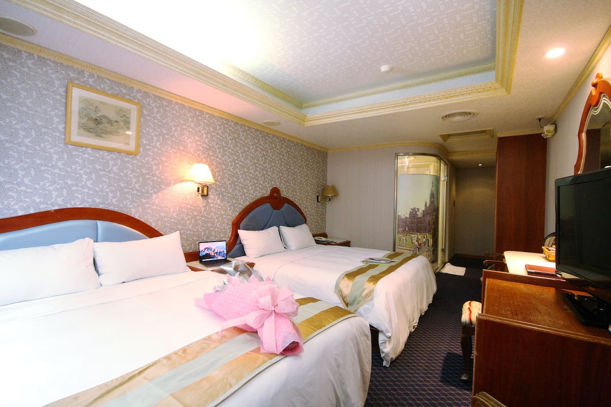 Habitación de hotel con dos camas, un televisor, un escritorio y un baño visible a través de una puerta abierta.