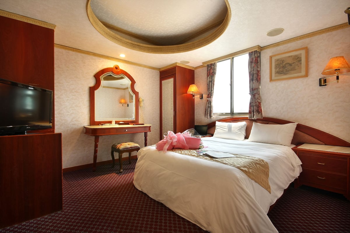 Habitación de hotel con una cama grande, televisor, escritorio con silla, espejo y ventana con cortinas.