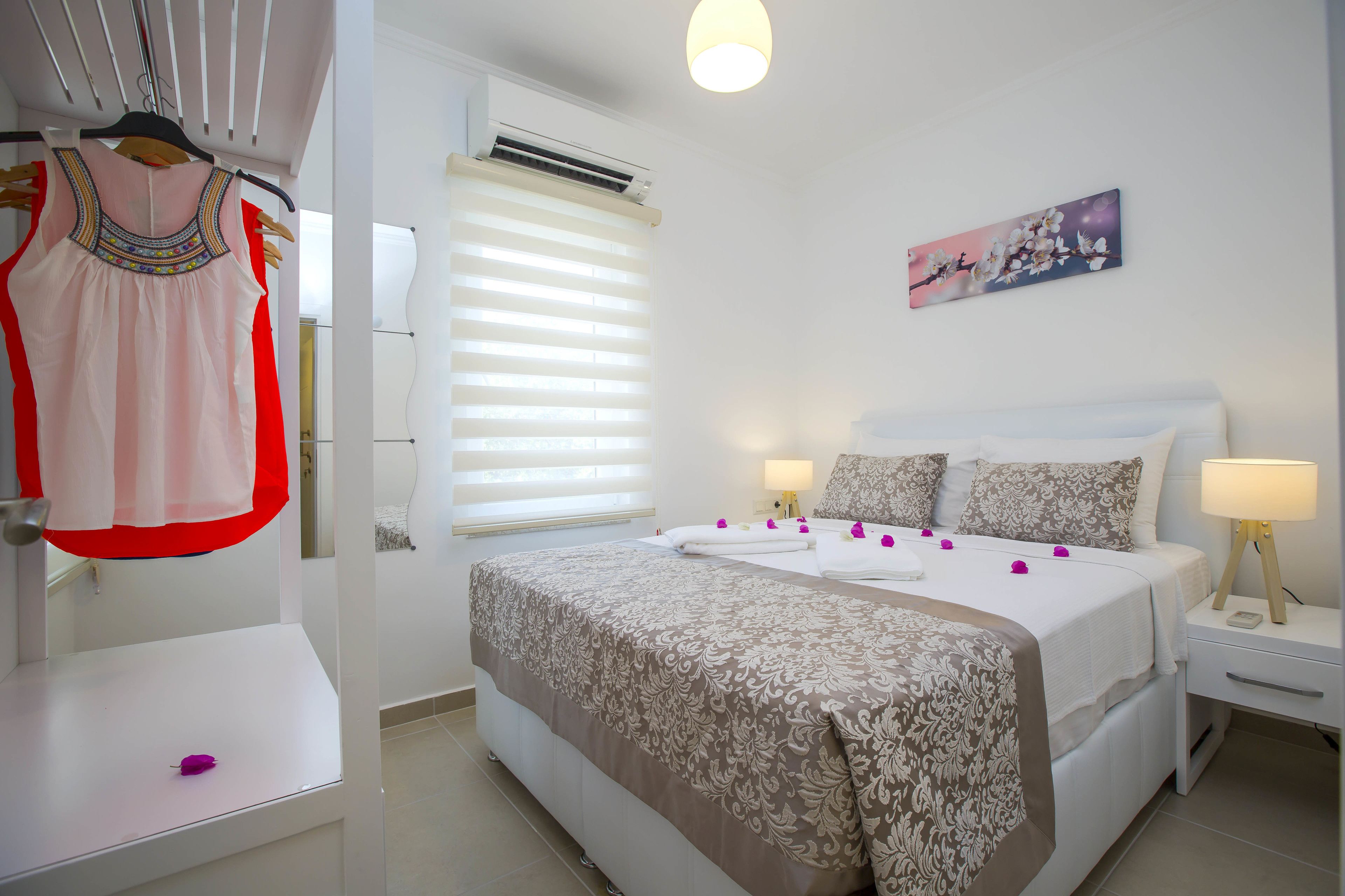 Une chambre d’hôtel comprenant un lit, deux tables de chevet, une commode, une fenêtre avec stores, un climatiseur fixé au mur et un porte-vêtements suspendu.