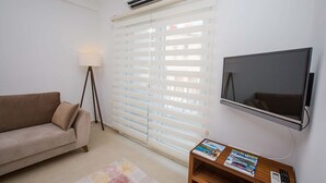Una sala de estar con un sofá color beige, una lámpara de pie, un televisor montado en la pared y una ventana con persianas.