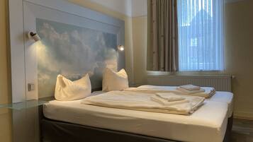 Un dormitorio con cama, almohadas, una mesita de noche de cristal, cabecero con un mural de nubes, ventana con cortinas y radiador.