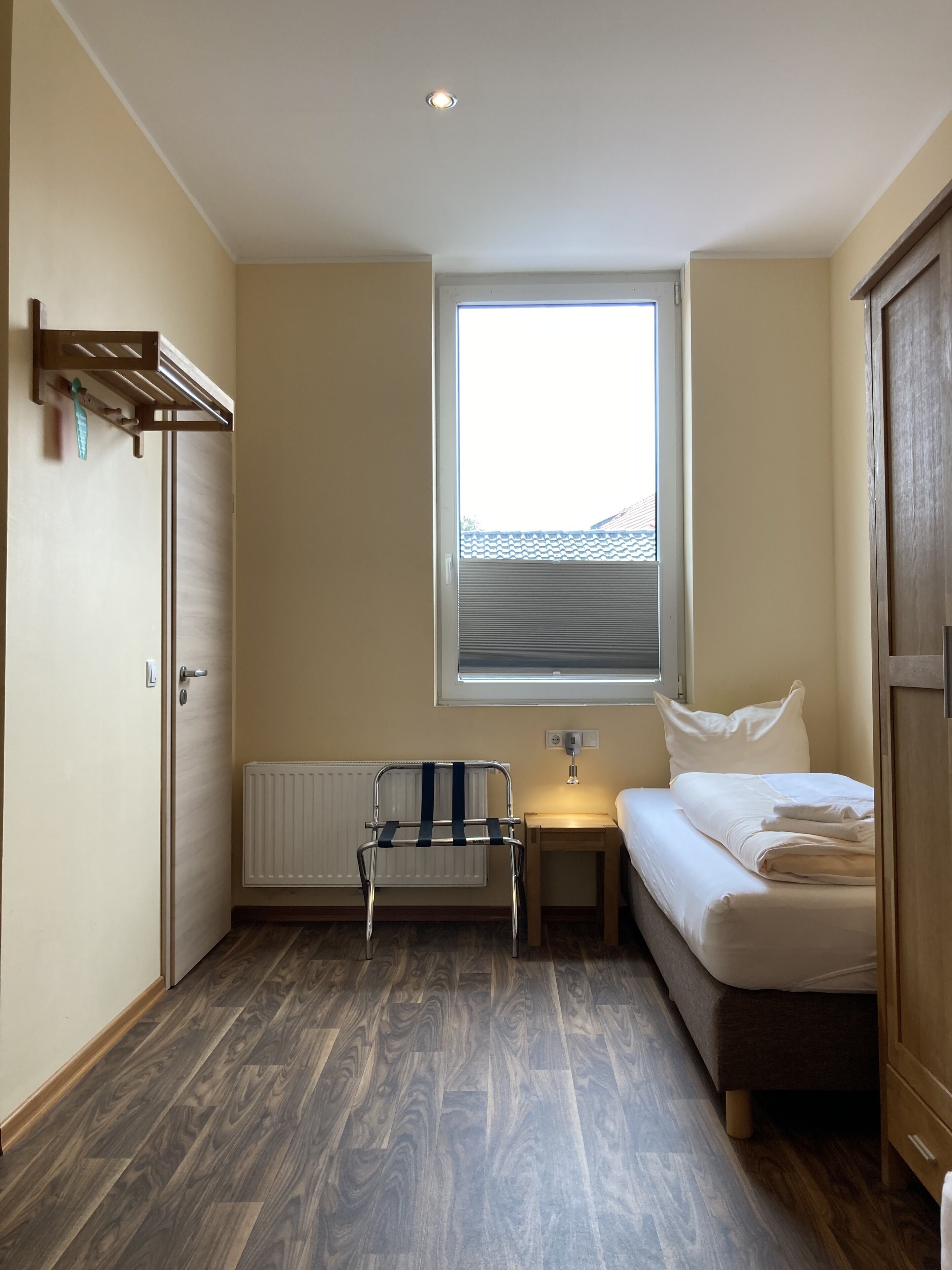 A small, well-lit room with a wooden floor, a single bed, a nightstand, a radiator, a window with blinds, a radiator cover, and a door.