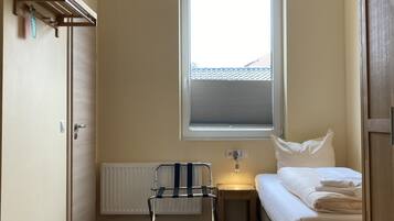 A small, well-lit room with a wooden floor, a single bed, a nightstand, a radiator, a window with blinds, a radiator cover, and a door.
