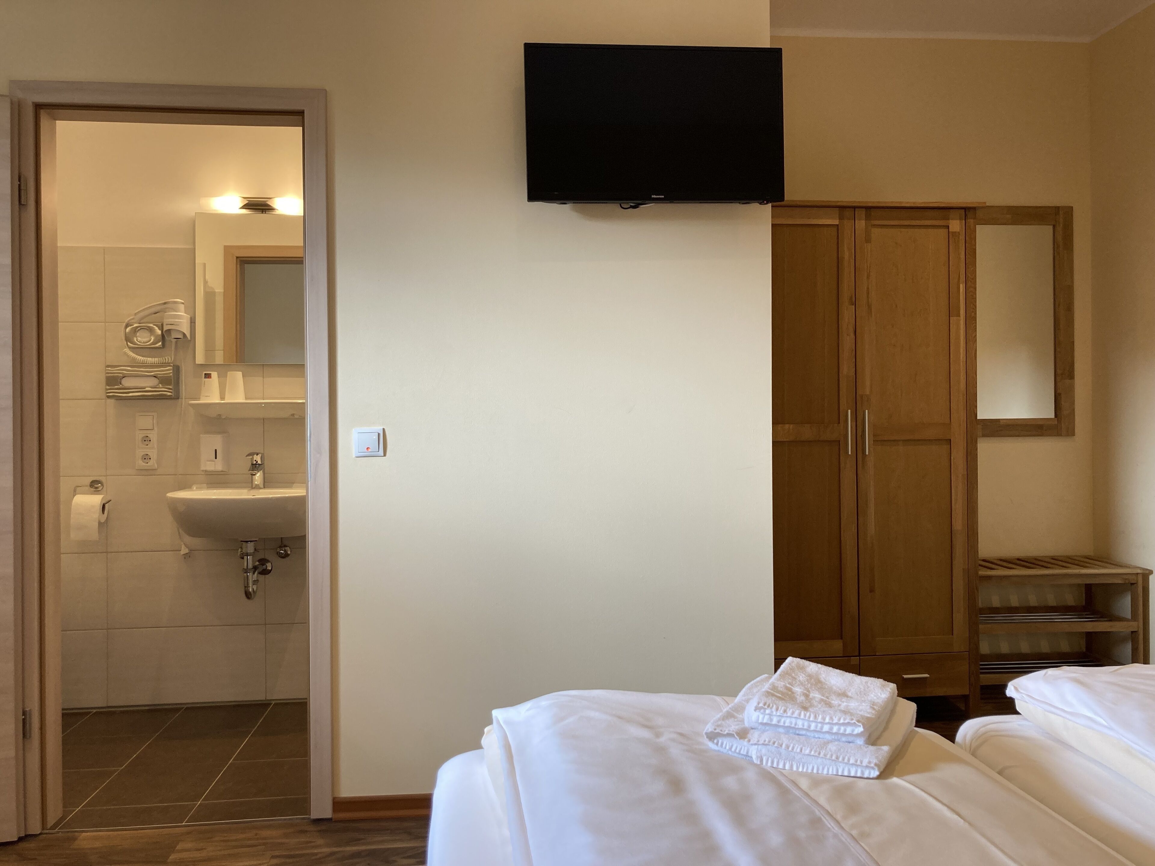 A hotel room with a bed, a TV mounted on the wall, a wooden wardrobe, and a bathroom visible through an open door.