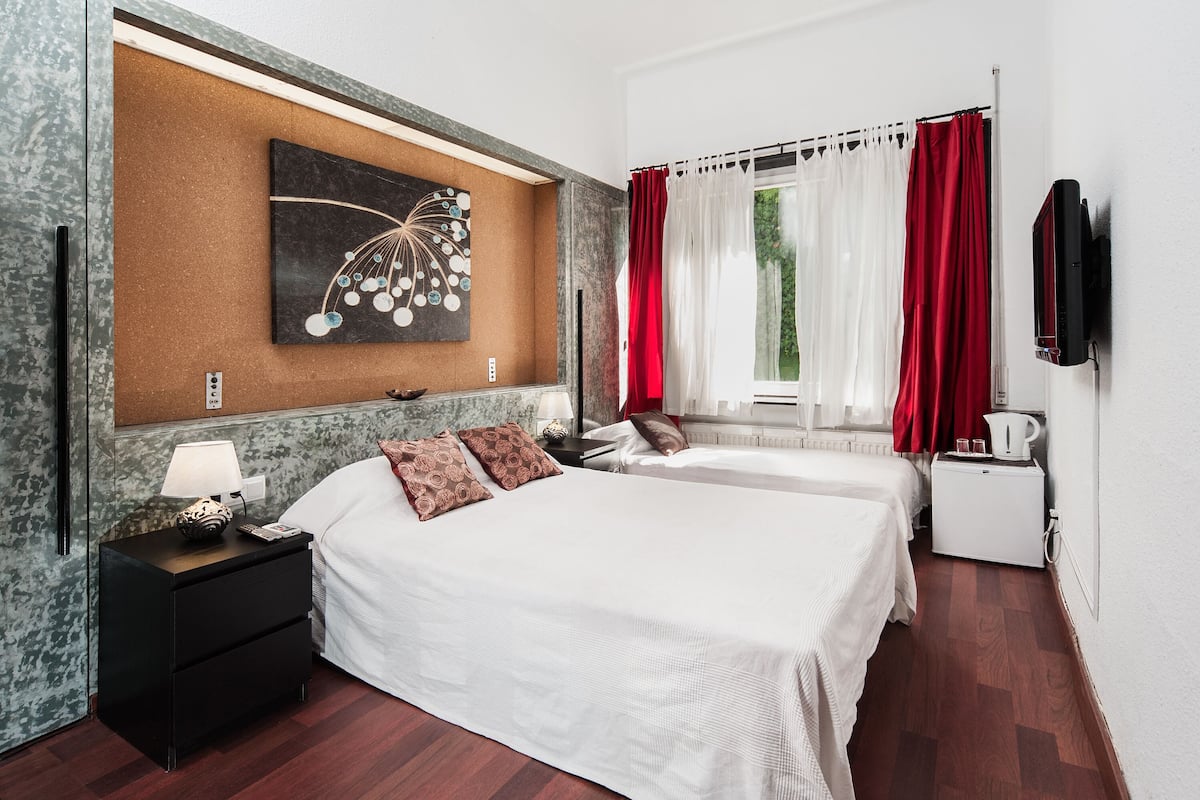 Una habitación de hotel con una cama grande, una mesita de noche con lámpara, un cuadro en la pared y una ventana con cortinas rojas.