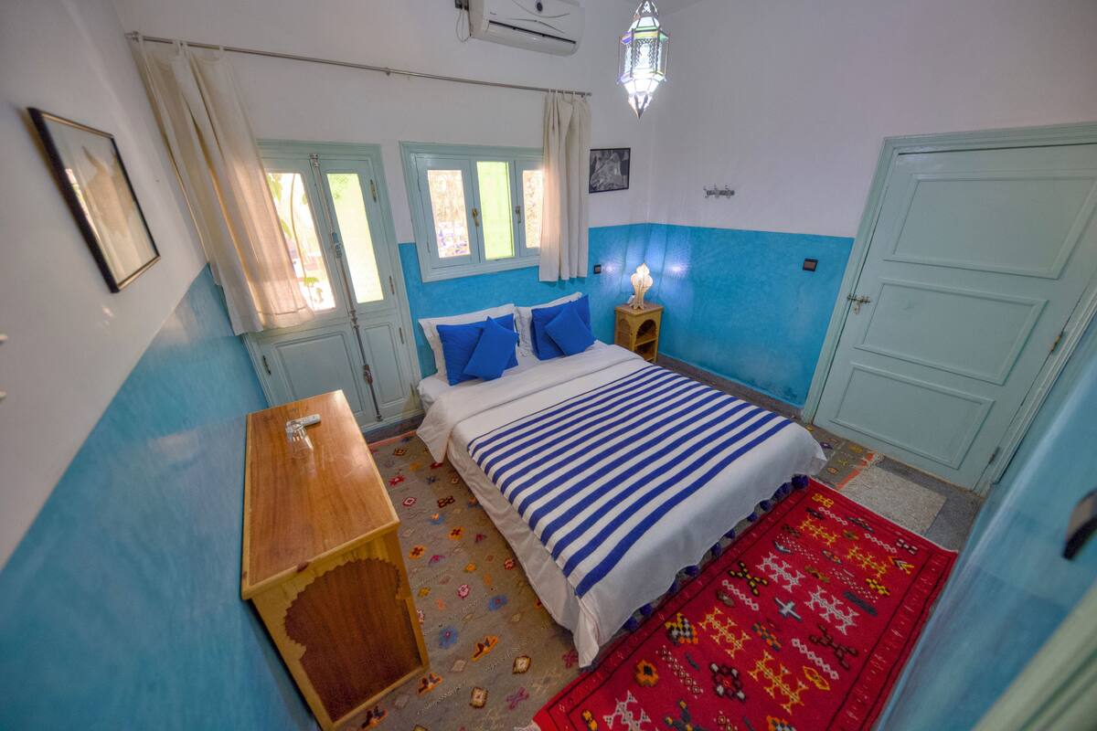 A bedroom with a bed, a wooden nightstand, a red rug, a window with curtains, and a wall-mounted air conditioner.