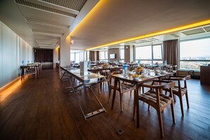 Un comedor amplio con varias mesas y sillas, ventanas grandes que ofrecen vista a la ciudad y un diseño de techo moderno.