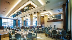 Un amplio salón de banquetes con varias mesas redondas y sillas, ventanas grandes y una lámpara moderna.