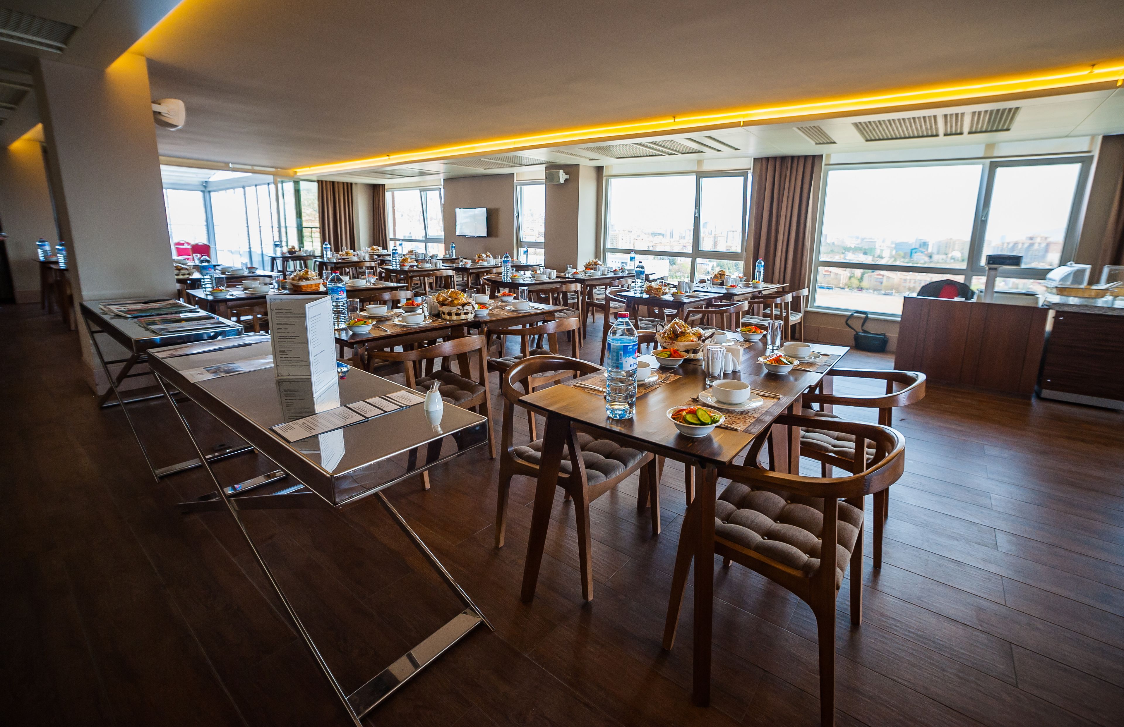 Un amplio comedor de hotel con múltiples mesas y sillas, ventanas grandes que ofrecen vistas a la ciudad y un diseño interior moderno.