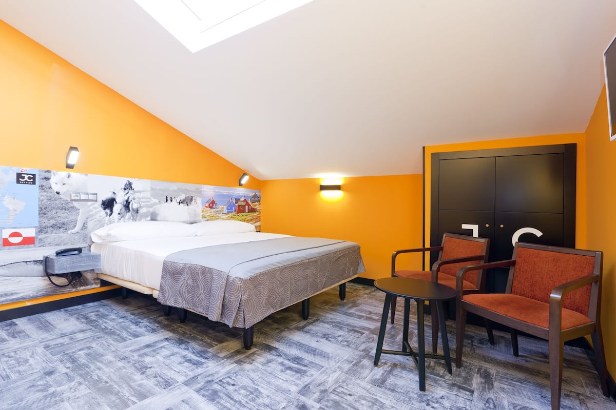 A hotel room with a bed, two chairs, a desk, and a large window.