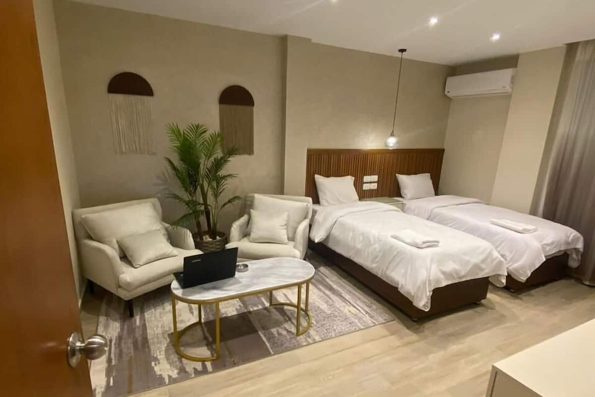 Quarto de hotel com sofá, duas camas individuais, uma mesinha e um vaso de planta.