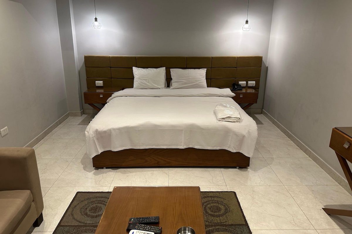 Cama bem arrumada com lençóis brancos, duas mesas de cabeceira com luminárias, cabeceira de madeira e uma pequena mesa com controles remotos em um quarto de hotel.