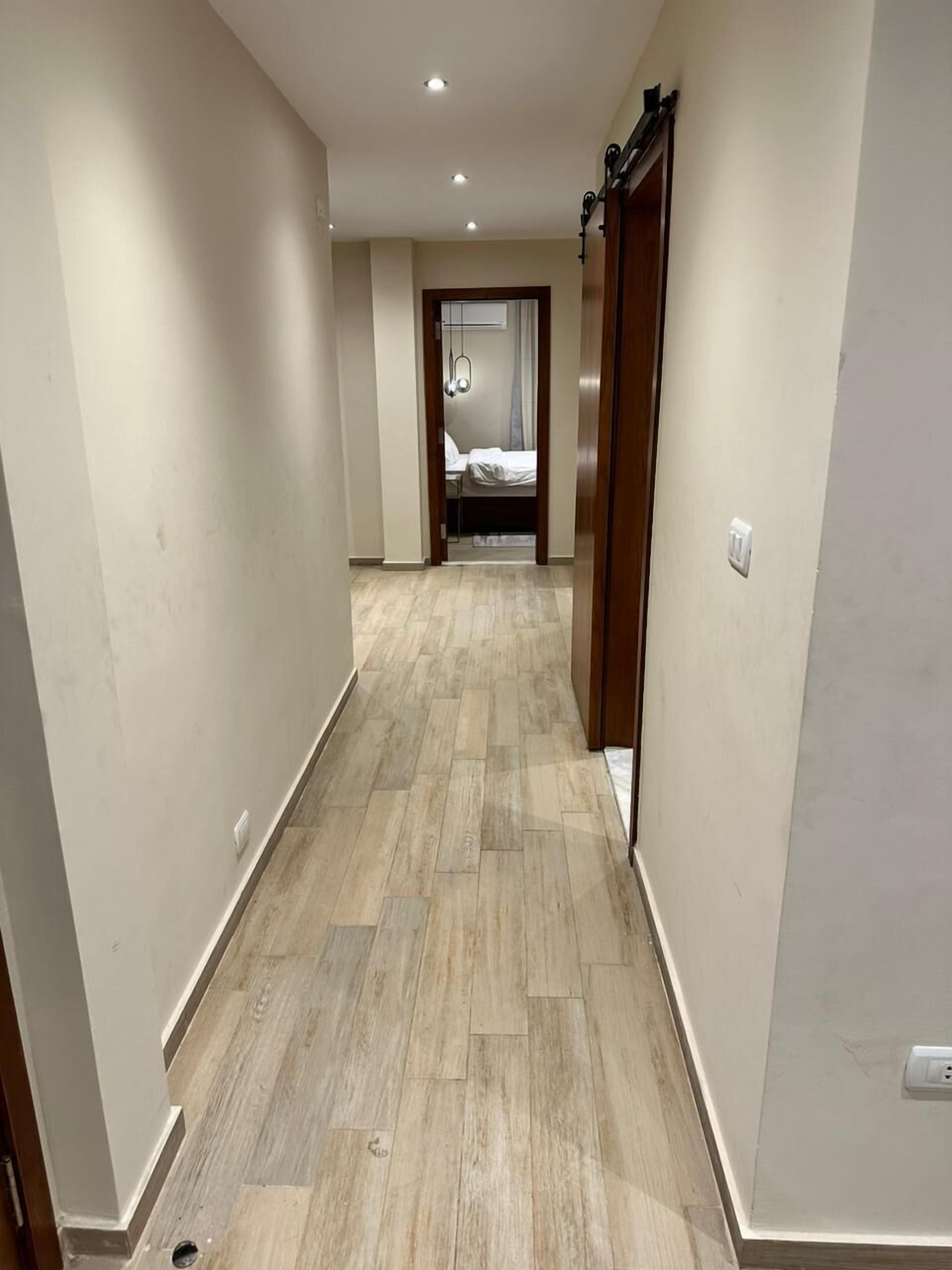 Un pasillo estrecho con suelo de madera y paredes blancas, que conduce a un dormitorio con cama y ventana.