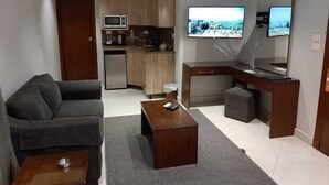 A compact living space with a sofa, a coffee table, a desk with a computer, a TV, and a kitchenette. - Nakhil Residence (Giza)