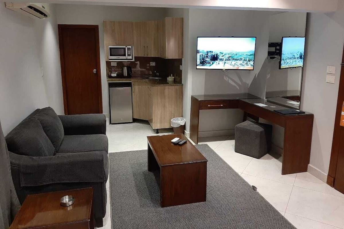 Espaço compacto com sofá, mesa de centro, escrivaninha com computador, televisão e pequeno espaço de cozinha.